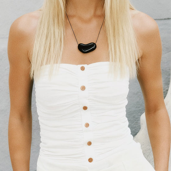 Woman wearing a white dress with buttons and a black heart-shaped necklace against a light gray background.