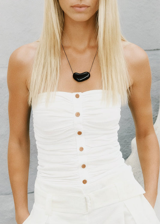 Woman wearing a white dress with buttons and a black heart-shaped necklace against a light gray background.