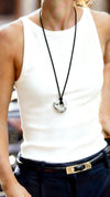 Woman wearing a white tank top and navy pants with sunglasses and a rope necklace with a silver near-oval pendant.