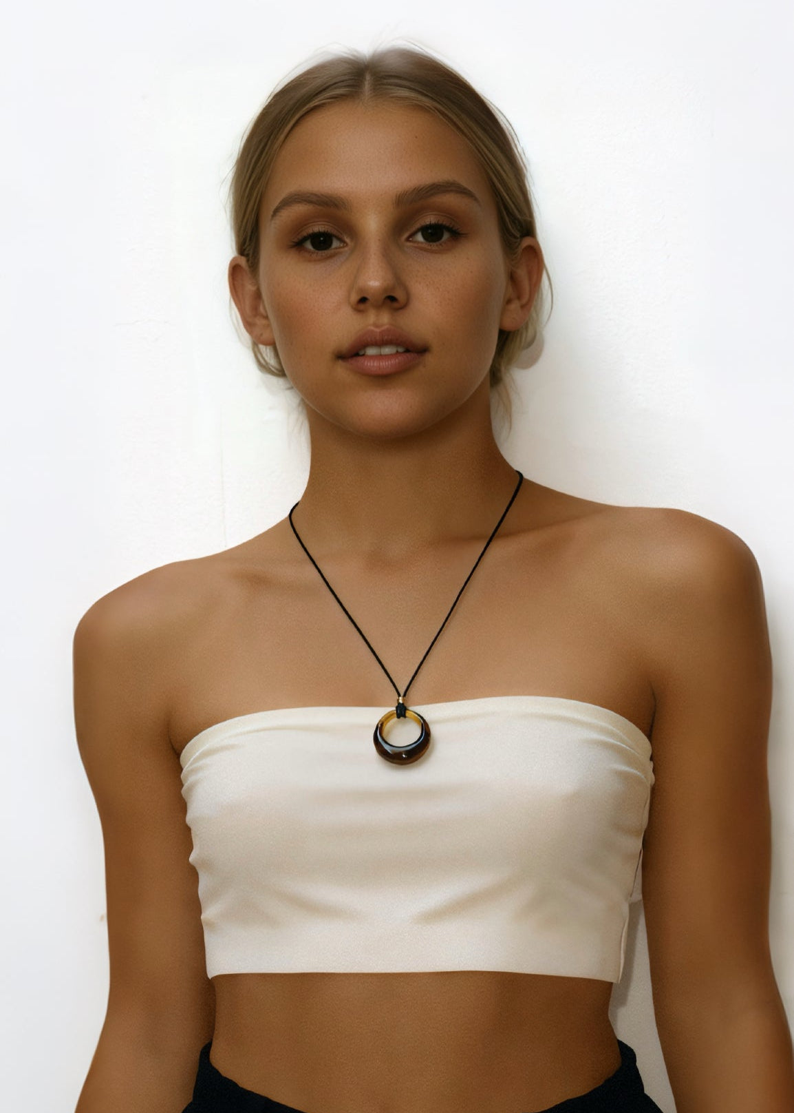 Woman wearing a white strapless top and black skirt with a amber circle pendant cord necklace on a white background