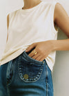 Person wearing a white sleeveless top and blue jeans and a gold ring with a neutral background