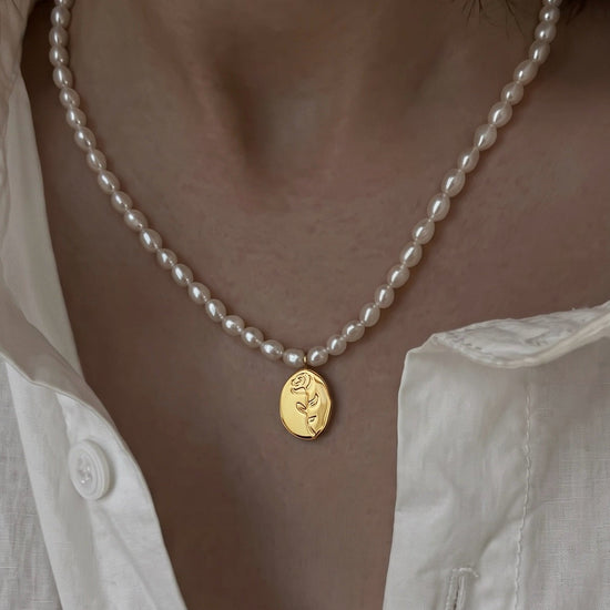 A close-up of a person wearing a delicate white pearl necklace with a small oval gold pendant featuring an engraved rose, styled with an open white linen shirt.
