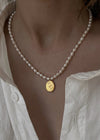 A close-up of a person wearing a delicate white pearl necklace with a small oval gold pendant featuring an engraved rose, styled with an open white linen shirt.