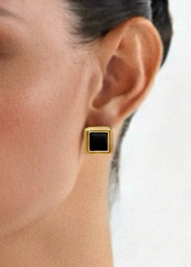 Close-up of an ear wearing a gold earring with a black square stone on a white background