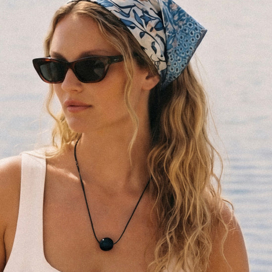 Woman wearing a black bead pendant on a black cord, a white outfit with sunglasses and a headscarf by a waterfront.