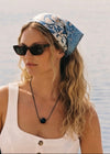 Woman wearing a black bead pendant on a black cord, a white outfit with sunglasses and a headscarf by a waterfront.