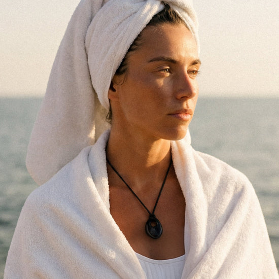 Woman wearing a black cord necklace and wrapped in a white towel by the ocean