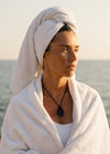 Woman wearing a black cord necklace and wrapped in a white towel by the ocean